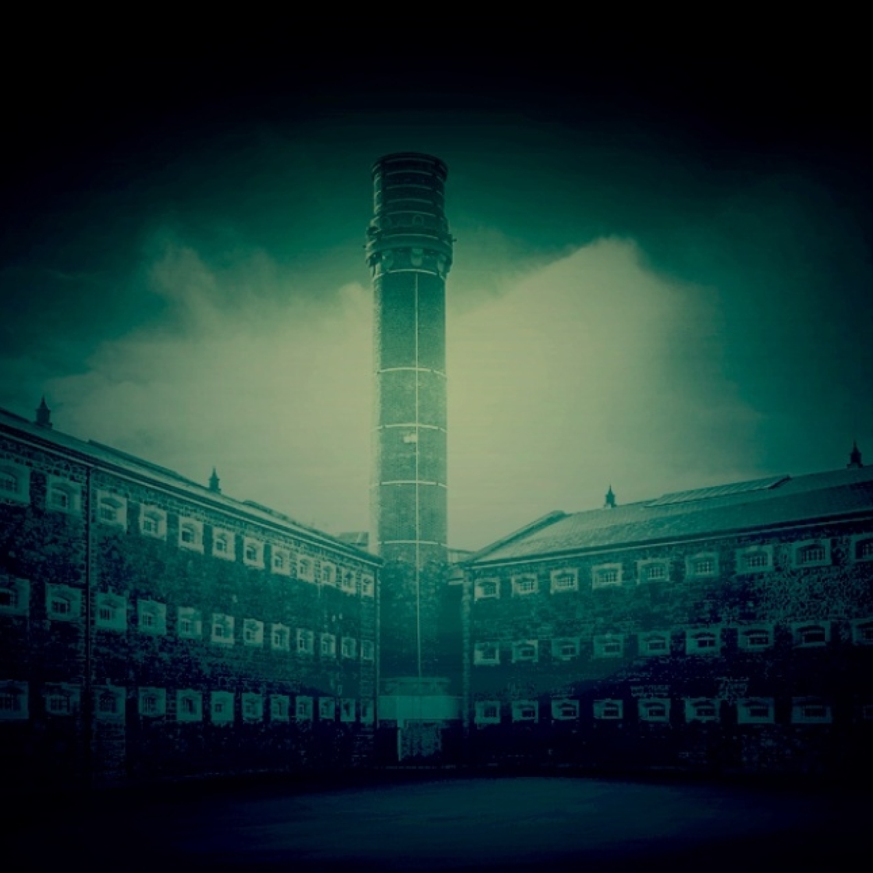 A green-blue tinted image of a tall, external brick tower overlooking three-storey Y-shaped brick prison wings containing dozens of small windows